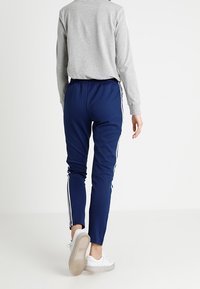 Marineblauwe sportbroek met witte zijstrepen, elastische tailleband en een slim fit, gecombineerd met een grijs langlopenshirt en witte sneakers.
