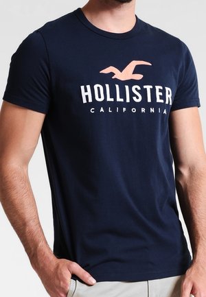 Mężczyzna noszący granatowy t-shirt Hollister California z różowym logo mewy, ręka w kieszeni i beżowe spodnie.