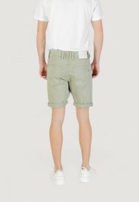 Pantalones cortos de mezclilla verde con puños enrollados, cinco bolsillos y un parche de cuero con marca en la cinturilla trasera. Llevados con una camiseta blanca y zapatillas.
