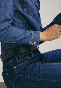 Personne assise portant une chemise en jean bleu et un jean avec une ceinture noire, les mains positionnées près de la taille, sur un fond uni clair.