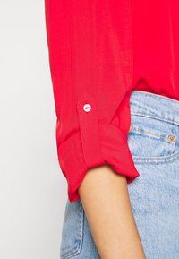 Rotes Langarmhemd mit aufgerolltem und oberhalb des Ellbogens geknöpftem Ärmel, kombiniert mit hellblauen Jeans.