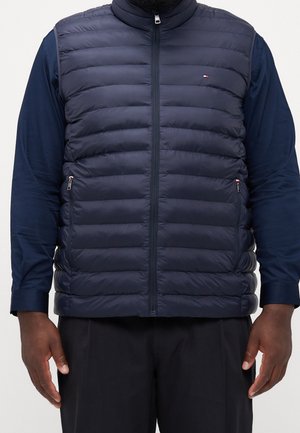 Uomo che indossa un gilet trapuntato blu navy con zip sopra una maglia a maniche lunghe blu navy e pantaloni neri, su uno sfondo semplice.