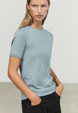 Jeune femme aux cheveux blonds et raides portant une chemise bleu clair à manches courtes et un pantalon noir, les mains dans les poches.