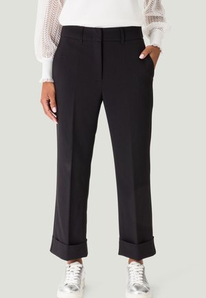 Frau trägt schwarze gerade geschnittene Hosen mit Umschlag, weiße Bluse mit Spitzenärmeln und silberne Turnschuhe, steht mit einer Hand in der Tasche.