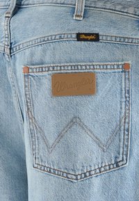 Τσέπη στην πλάτη από γαλάζιο τζιν με ραμμένο σχέδιο "W" σε ταμπά χρώμα, καφέ δερμάτινο ταμπελάκι και μικρή μαύρη υφασμάτινη ετικέτα, και τα δύο με την ένδειξη Wrangler.