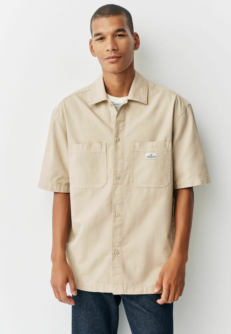 Un homme portant une chemise beige à manches courtes avec deux poches avant et un jean bleu foncé, se tenant devant un fond blanc.