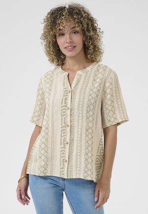 Blouse beige à manches courtes avec des motifs géométriques verts, devant boutonné et col rond. Fabriquée en tissu léger pour plus de confort.