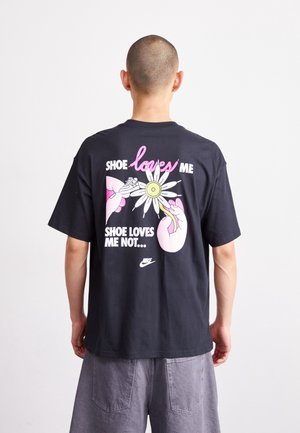 Persoon met geschoren hoofd die een zwart shirt draagt met een roze en wit beeld van handen die madeliefblaadjes plukken, met de tekst "Schoen houdt van mij, schoen houdt niet van mij."