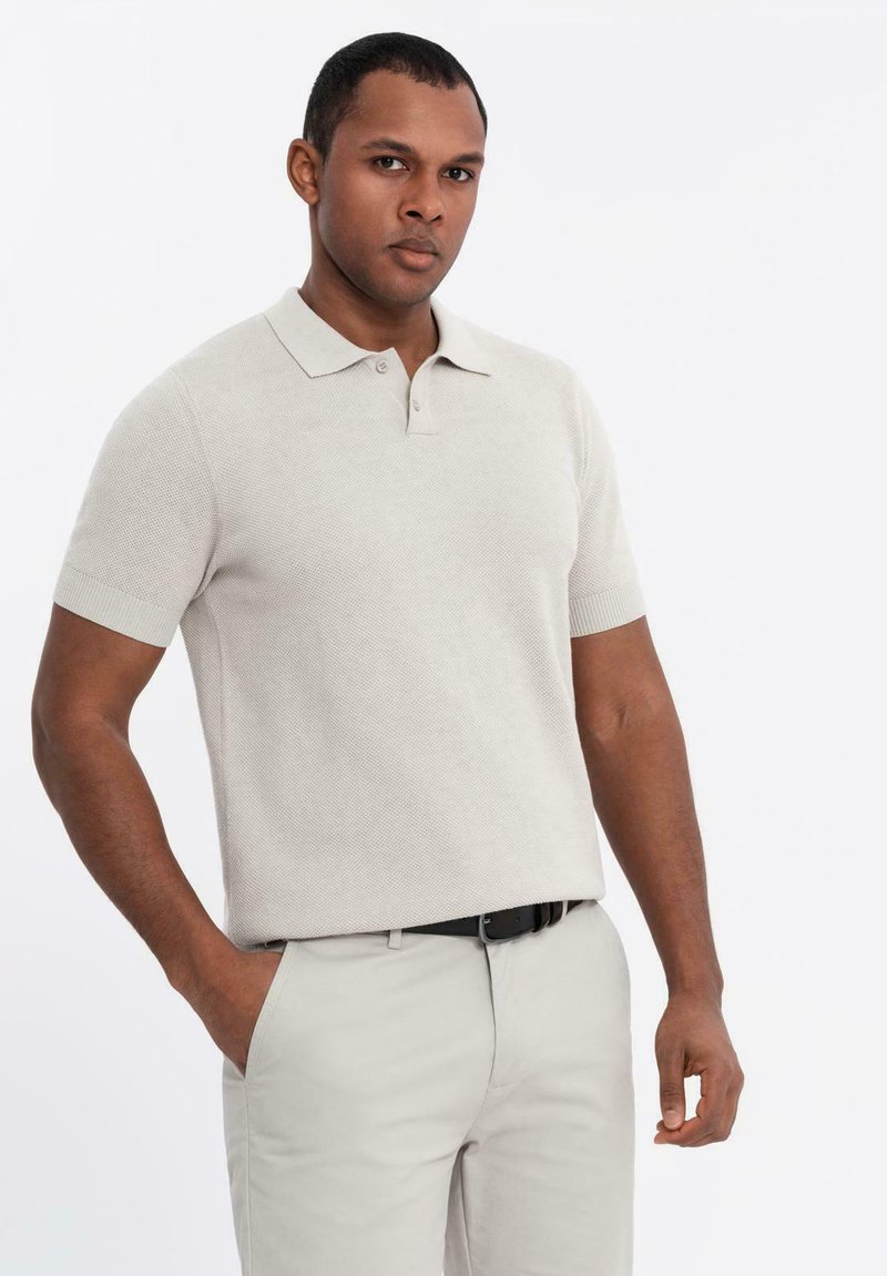 Beige strikket polo med korte ærmer, struktureret stof, tre-knaps lukning, båret med lyse bukser, stående foran en hvid baggrund.