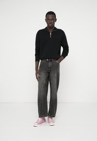 Vyras stovi su ranka kišenėje, vilkėdamas juodą užtrauktuku užsegamą megztinį, tamsiai mėlynus plačius džinsus, juodą diržą ir rožinius aukštus sportbačius ant balto fono.