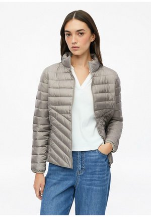 Femme portant une veste matelassée gris clair, un t-shirt blanc à col en V et un jean bleu, debout avec une main dans la poche sur fond blanc.