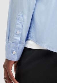 Polsino di una camicia a maniche lunghe blu chiaro con bottoni bianchi su una mano di carnagione scura, abbinato a pantaloni neri e una maglietta bianca visibile.
