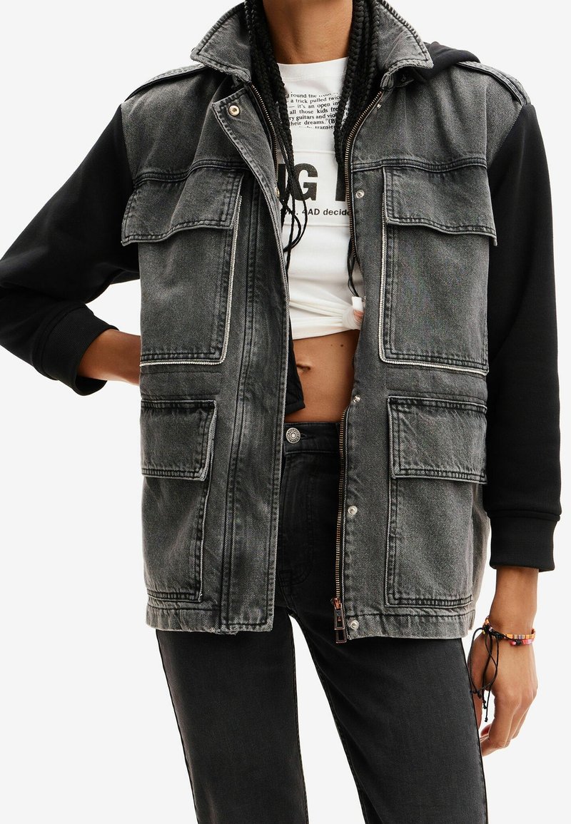 Personne portant une veste en jean noire et grise avec de grandes poches avant, des manches noires et un t-shirt graphique blanc court en dessous.