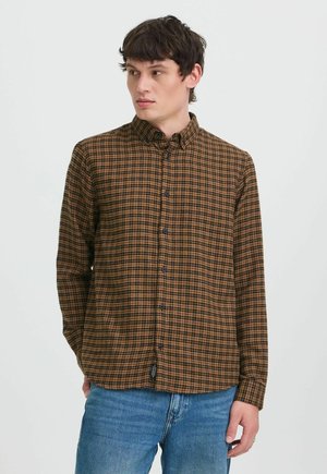 Jonge man met een bruine en zwarte geruite blouse met knopen en een blauwe spijkerbroek, kijkt naar links tegen een effen achtergrond.