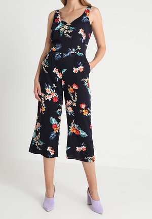 Un mono floral de color navy con escote en V, piernas anchas y tirantes. Presenta brillantes patrones florales en rojo, rosa y azul verdoso sobre un fondo oscuro.