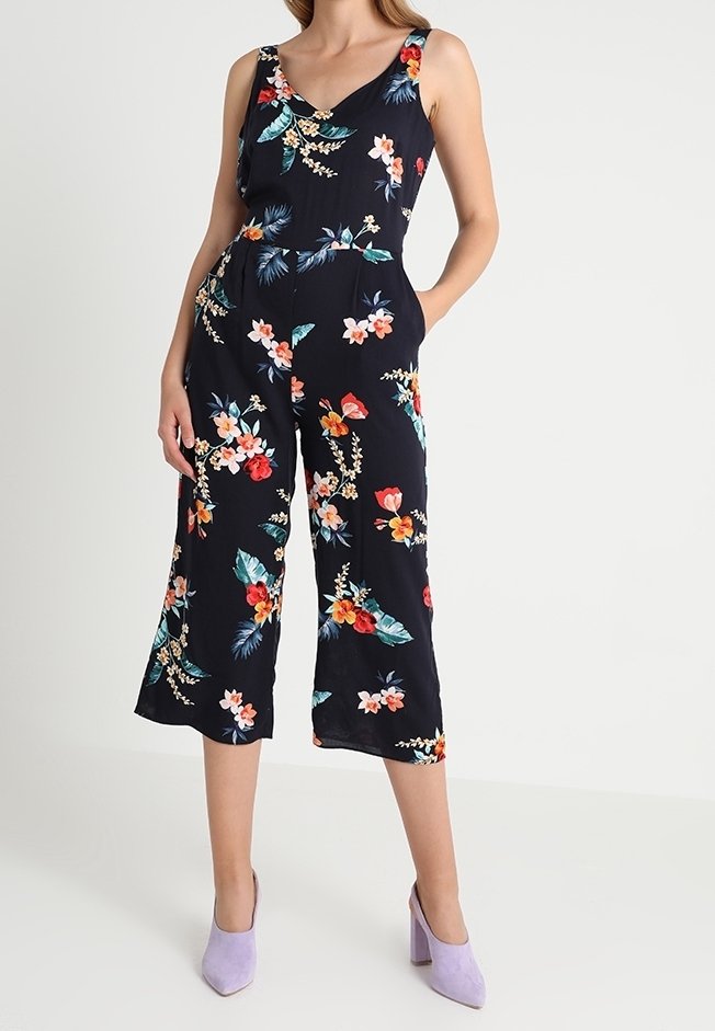Marinblå blommig jumpsuit med V-ringning, vida ben och axelband. Har klara blommönster i röd, rosa och teal på en mörk bakgrund.