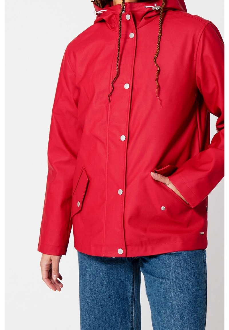 Chaqueta impermeable roja con capucha, botones de presión, bolsillos laterales y cordón ajustable. Presenta una textura suave y un corte holgado.