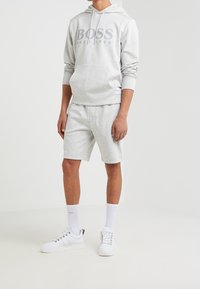 Homme portant un sweat à capuche Hugo Boss gris clair et un short assorti, des chaussettes blanches et des baskets blanches sur un fond blanc uni.