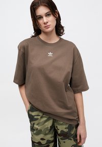 Tricou din bumbac maro, cu mâneci scurte și decolteu rotund. Are un logo alb Adidas pe piept. Asortat cu pantaloni cu model camuflat.
