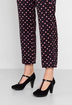 Beine, die schwarze Hosen mit rot-weißem Rautenmuster tragen, kombiniert mit schwarzen T-Riemchen High Heels auf einem hellen Boden.