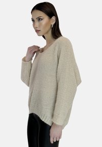 Beżowy sweter robiony na drutach z dekoltem w kształcie litery V, opadającymi ramionami i luźnym krojem. Tekstura jest miękka, z prążkowanymi detalami przy hemie i szerokimi rękawami.