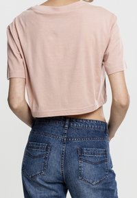 Persona che indossa una maglietta corta rosa chiaro e jeans a vita alta blu, mostrata di spalle contro uno sfondo semplice.