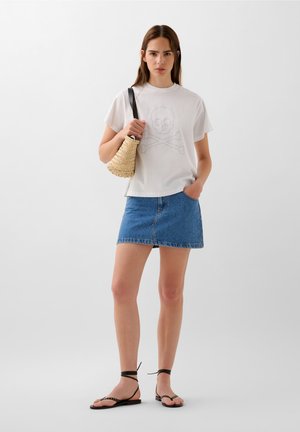 T-shirt bianca in cotone con stampa di teschio e ossa incrociate, abbinata a una mini gonna di jeans blu, sandali neri con cinturini e una borsa intrecciata.