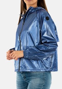 Glanzende blauwe lichtgewicht jas met een rits aan de voorkant, elastische manchetten en een capuchon. Bevat een logo patch op de linker mouw.