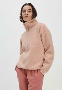 Heller rosa Fleece-Pullover mit hohem Kragen, Viertelreißverschluss und lockerer Passform. Weicher Stoff, diagonale Nahtdetails, kombiniert mit passenden rosa Hosen.