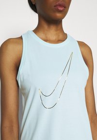 Vaaleansininen hihaton toppi pehmeästä kankaasta, jossa on kullanvärinen Nike-logo edessä. Sileä pinta, pyöreä kaula-aukko, rento malli.