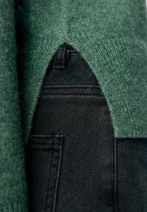 Detalle de un suéter de punto verde con un dobladillo en forma de V superpuesto sobre la parte superior de unos jeans de mezclilla negros.