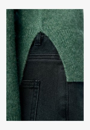 Pogled od blizu na zeleni pleteni pulover z V-oblikovanim robom, ki prekriva vrh črnih kavbojskih hlač.