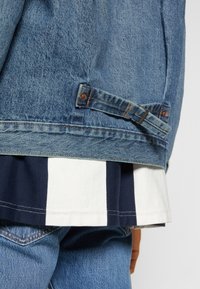 Jeansjacka i ljusblått med en texturerad yta. Inkluderar ett justerbart band på sidan och är lager på lager över en mörk- och vitrandig skjorta.