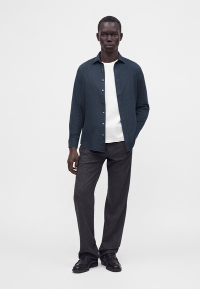 Homme portant une chemise bleu foncé à boutons par-dessus un t-shirt blanc, avec un pantalon gris foncé et des chaussures noires, debout devant un fond clair uni.
