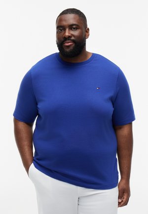 Άνδρας με γενειάδα που φοράει μπλε κοντομάνικο πουκάμισο και λευκό παντελόνι, στέκεται με τα χέρια στις τσέπες σε λευκό φόντο.