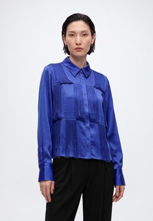 Femme aux cheveux courts et foncés portant un chemisier bleu vif à franges à manches longues et un pantalon noir, debout devant un fond blanc uni.