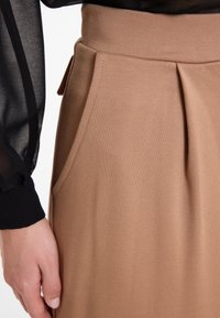 Bruine geplooide rok van textuur geweven stof, met zijzakken en een gladde tailleband. Een zwart doorschijnend top met lange mouwen is gedeeltelijk zichtbaar.
