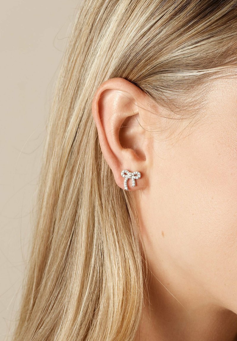 Silverörhängen formade som en båge, prydda med små klara rhinestones, synliga mot en ljusbeige bakgrund och bärs i ett hål i örat.