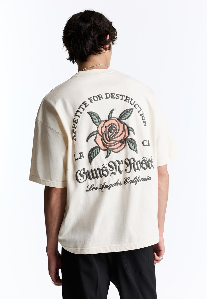 Personne aux cheveux foncés portant un t-shirt blanc Guns N’ Roses avec une grande rose et un texte dans le dos, associé à un pantalon noir.