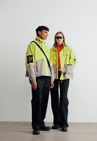 Δύο μοντέλα φορούν κίτρινες και γκρι μπουφάν, μαύρο παντελόνι και μαύρα παπούτσια· το ένα φοράει γυαλιά ηλίου, στέκεται μπροστά σε απλό λευκό φόντο.