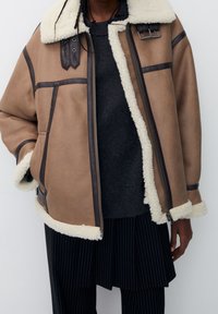 Giacca in shearling marrone con colletto e fodera in shearling crema, accentuata da piping marrone scuro e hardware in metallo, dotata di chiusura con zip.