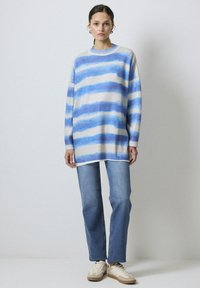 Oversized trui met blauw en witte strepen, geribbelde halslijn en lange mouwen, gecombineerd met lichtblauwe rechte jeans en casual sneakers.