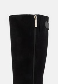 Botă din suede negru, de înălțime până la genunchi, cu o fermoar argintie și detalii de cusătură. Are o textură netedă și un accent cu curea pe lateral.