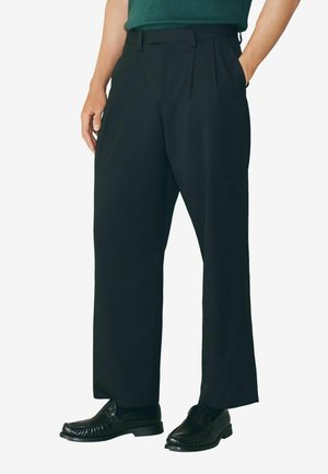 Schwarze, plissierte Hose mit geradem Bein, aus glattem Stoff gefertigt. Tragen mit schwarzen Slippern. Verfügt über Gürtelschlaufen und Seitentaschen.