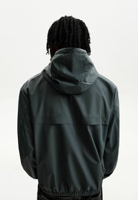 Person with braided hair wearing a dark gray hooded jacket, facing away from the camera against a plain white background.