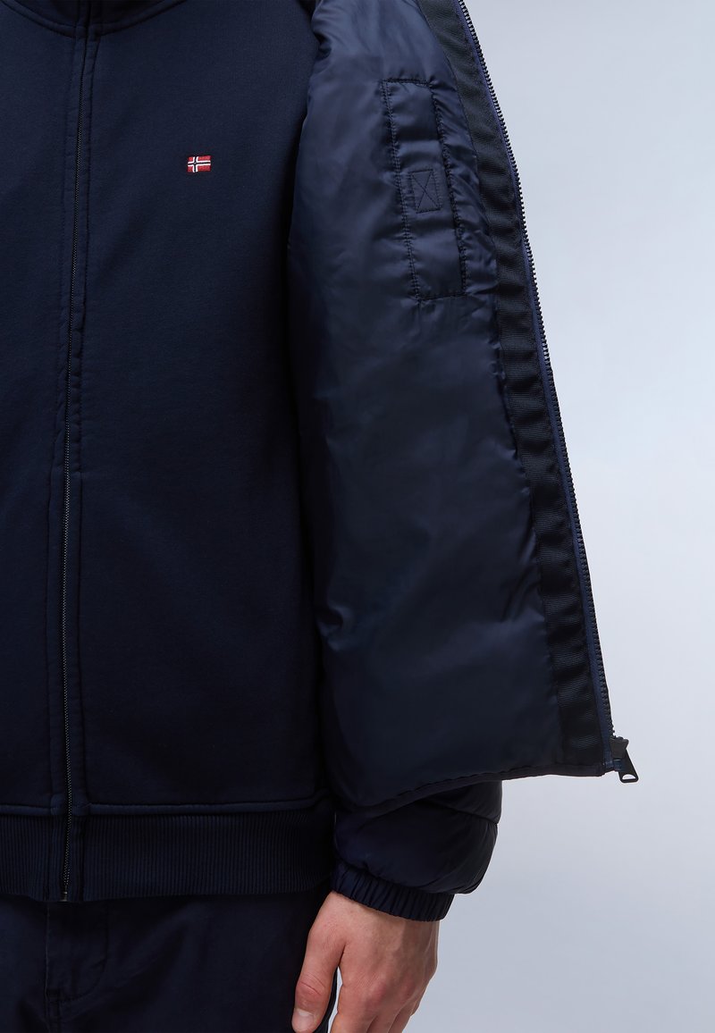 Granatowa bluza z zamkiem, z prążkowanymi mankietami i dołem. Posiada gładką, wyściełaną podszewkę oraz małe logo na piersi.