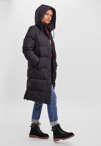 Manteau long matelassé noir avec capuche, poches latérales et fermeture éclair à l'avant. Porté par-dessus un jean blue et des bottes noires à lacets avec une semelle en caoutchouc.