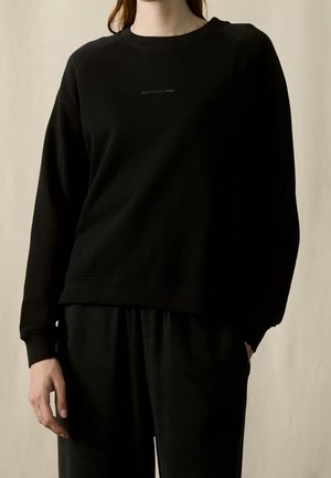 Personne portant un sweat-shirt noir uni à manches longues et col rond ainsi qu'un pantalon noir, debout avec une main dans la poche devant un fond neutre.