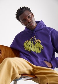 Paarse hoodie met een graffiti-stijl logo in geel, gecombineerd met tan broek. De stof lijkt zacht, met een ontspannen pasvorm en trekkoorden.