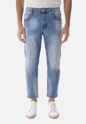 Uomo con jeans azzurri sbiaditi e sneakers bianche, in piedi con le braccia rilassate lungo i fianchi contro uno sfondo semplice.